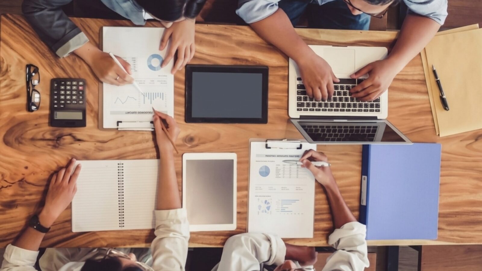 Vista dall'alto di quattro persone sedute a un tavolo di legno con laptop, tablet, documenti con grafici e quaderni