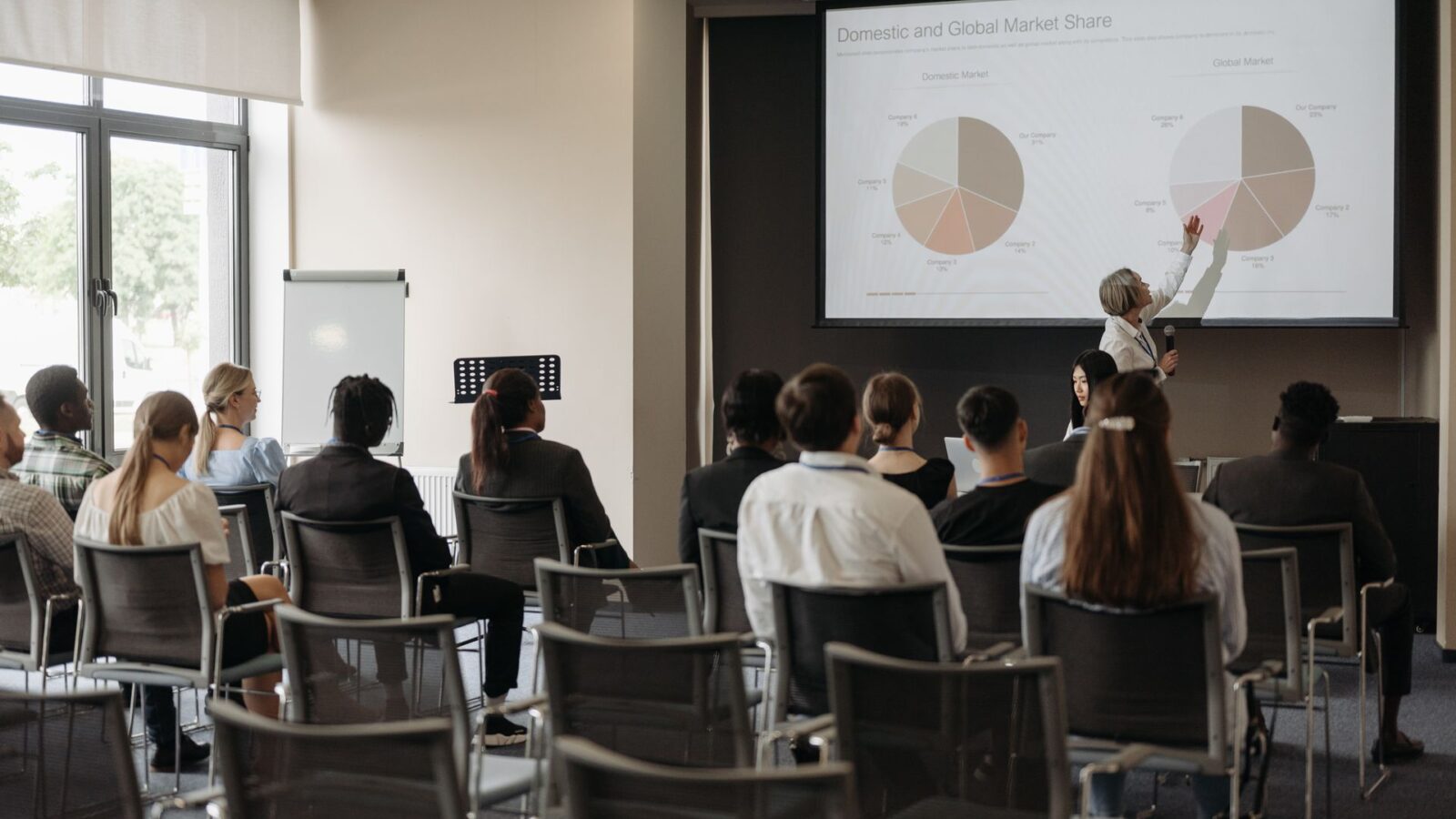 Relatrice indica lo schermo con la slide "Domestic and Global Market Share" davanti a una platea seduta in sala