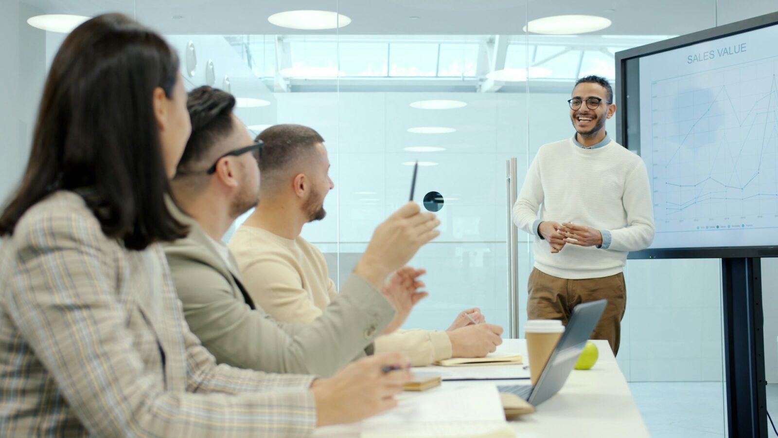 Un uomo sorridente presenta davanti a uno schermo con grafici e la scritta "SALES VALUE" a colleghi seduti al tavolo