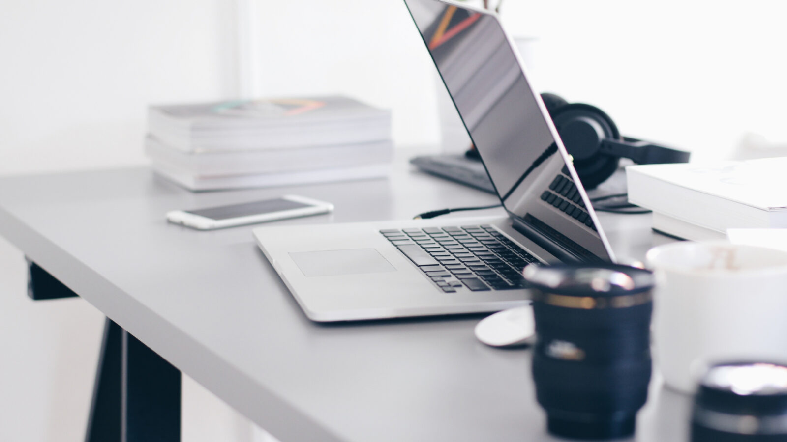Scrivania grigia con laptop aperto, smartphone, pila di libri, cuffie e un obiettivo fotografico