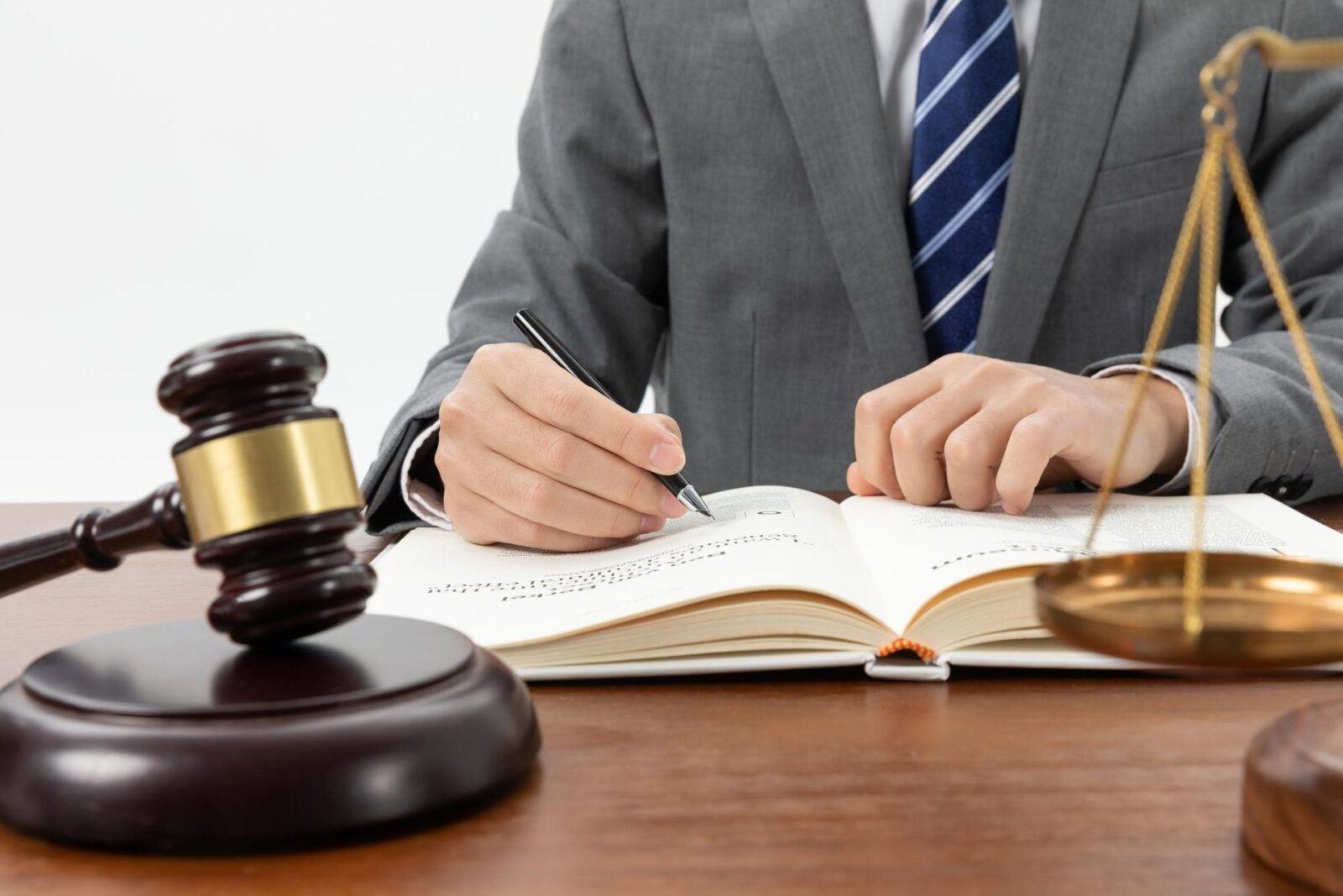 Uomo in giacca grigia e cravatta a righe blu scrive su un libro aperto tra un martelletto di legno e una bilancia dorata