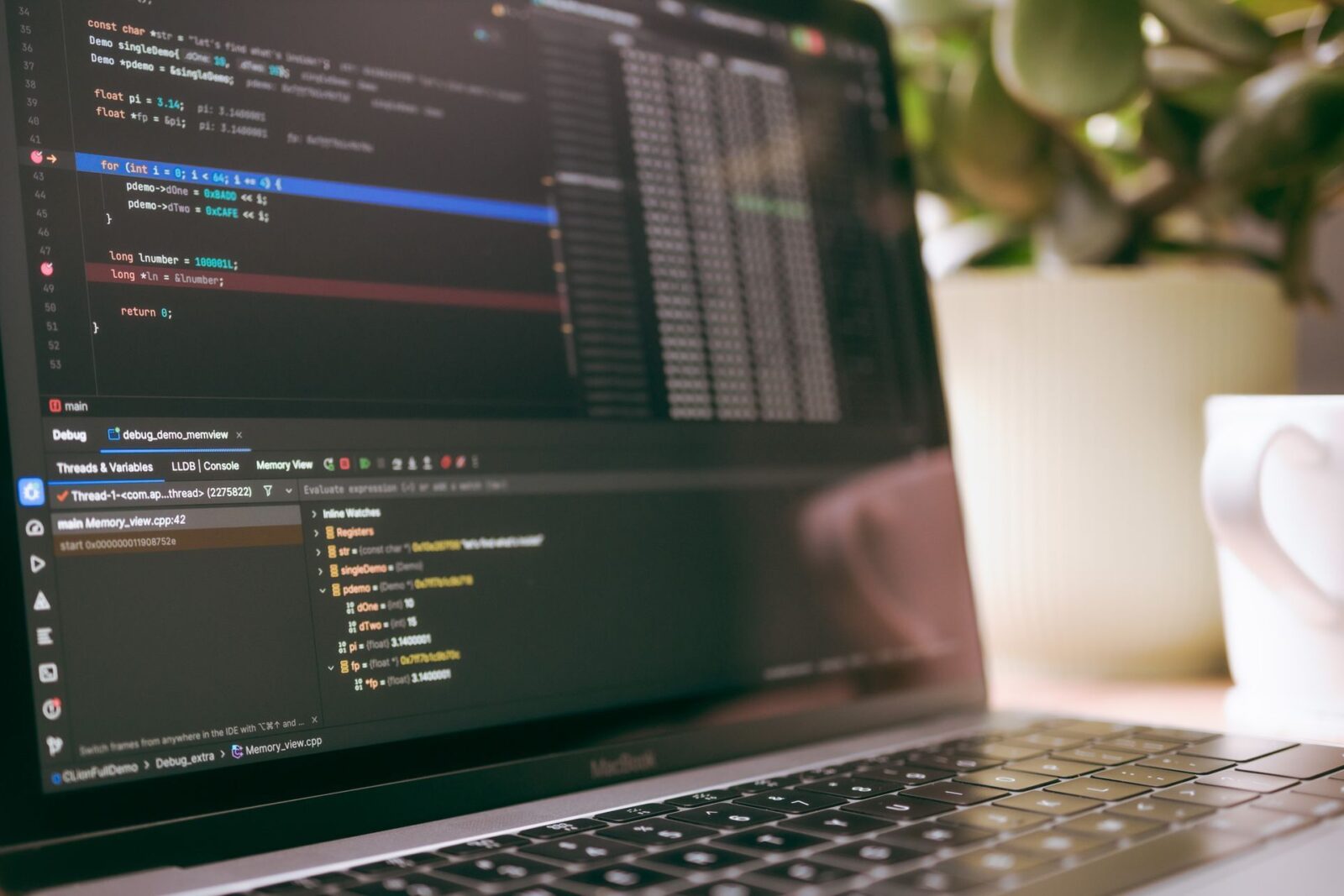 Schermo di un laptop con codice sorgente e pannello di debug, accanto a una pianta e a una tazza bianca sfocate