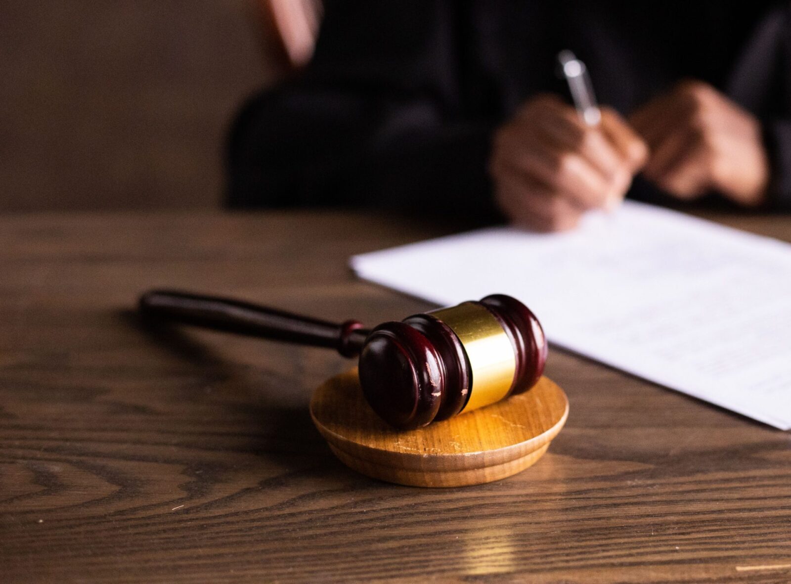 Martelletto da giudice su base circolare appoggiato a un tavolo di legno; sullo sfondo una persona firma un documento