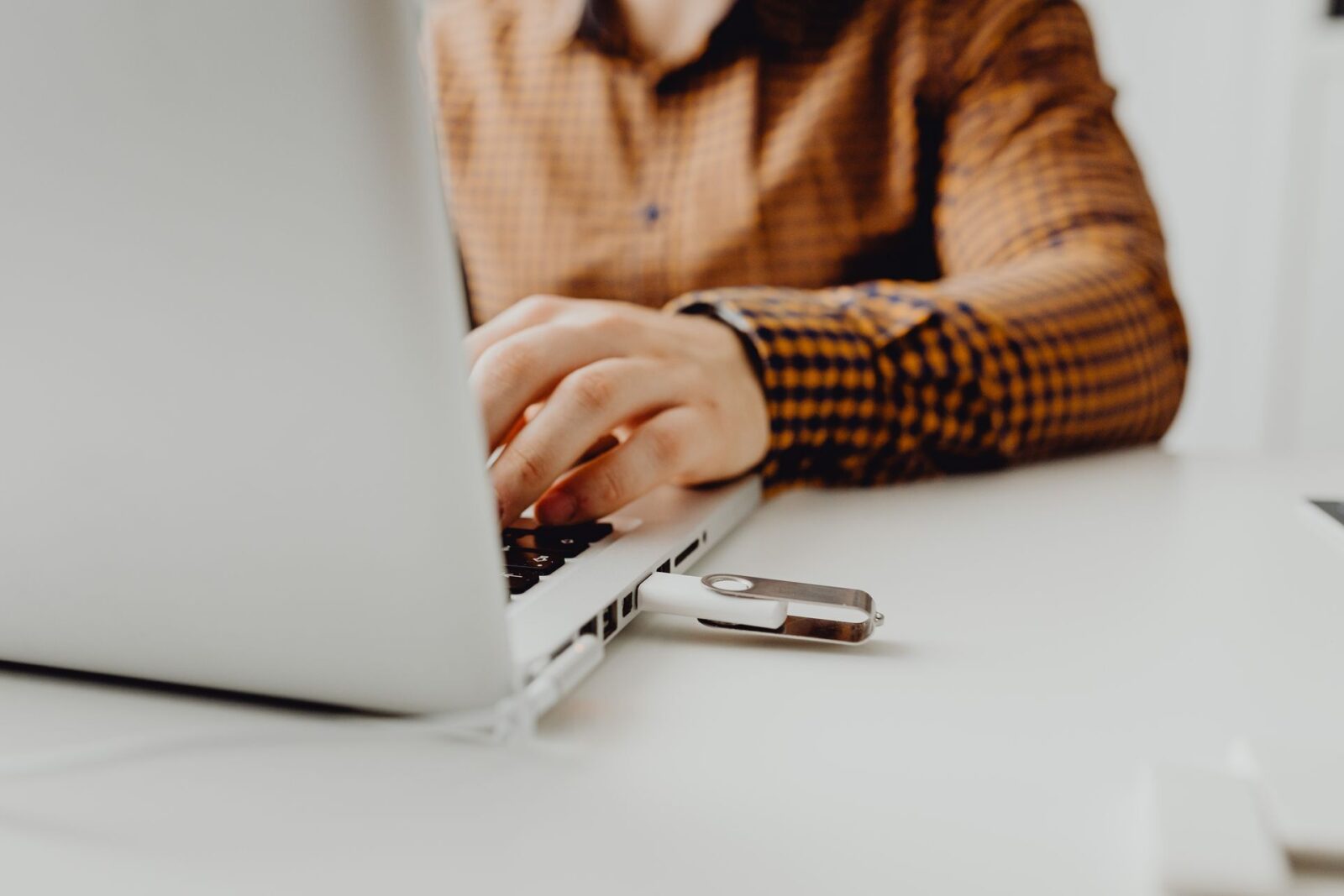 Mani di una persona in camicia a quadri digitano su un laptop grigio con una chiavetta USB inserita nella porta laterale