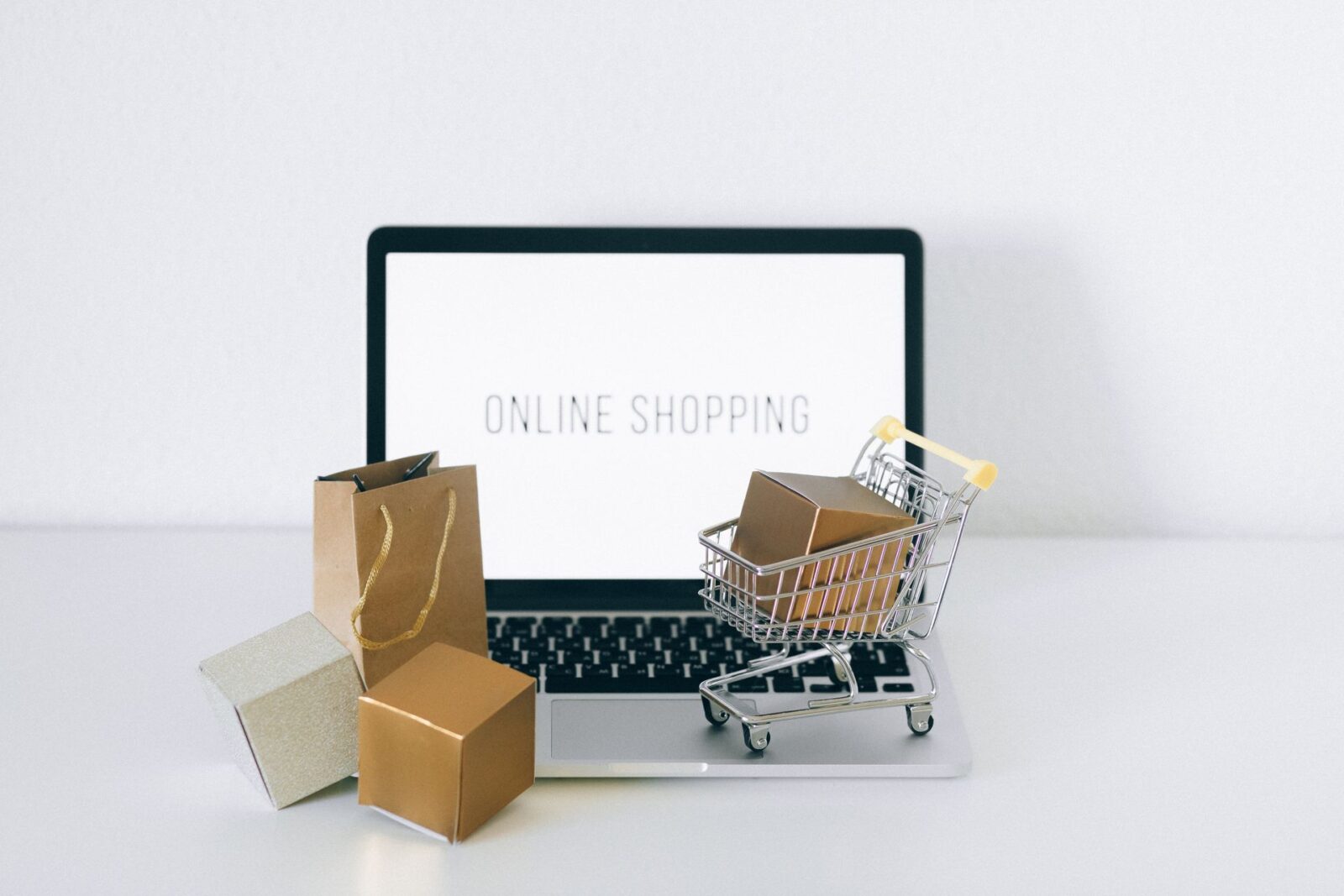 Laptop con la scritta ONLINE SHOPPING sullo schermo, davanti un carrello della spesa in miniatura, scatoline e sacchetto