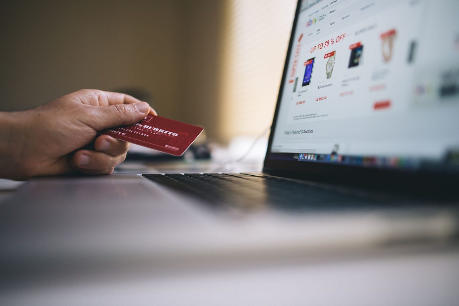 Mano che tiene una carta di credito rossa davanti a un portatile con un sito di acquisti e la scritta UP TO 70 % OFF