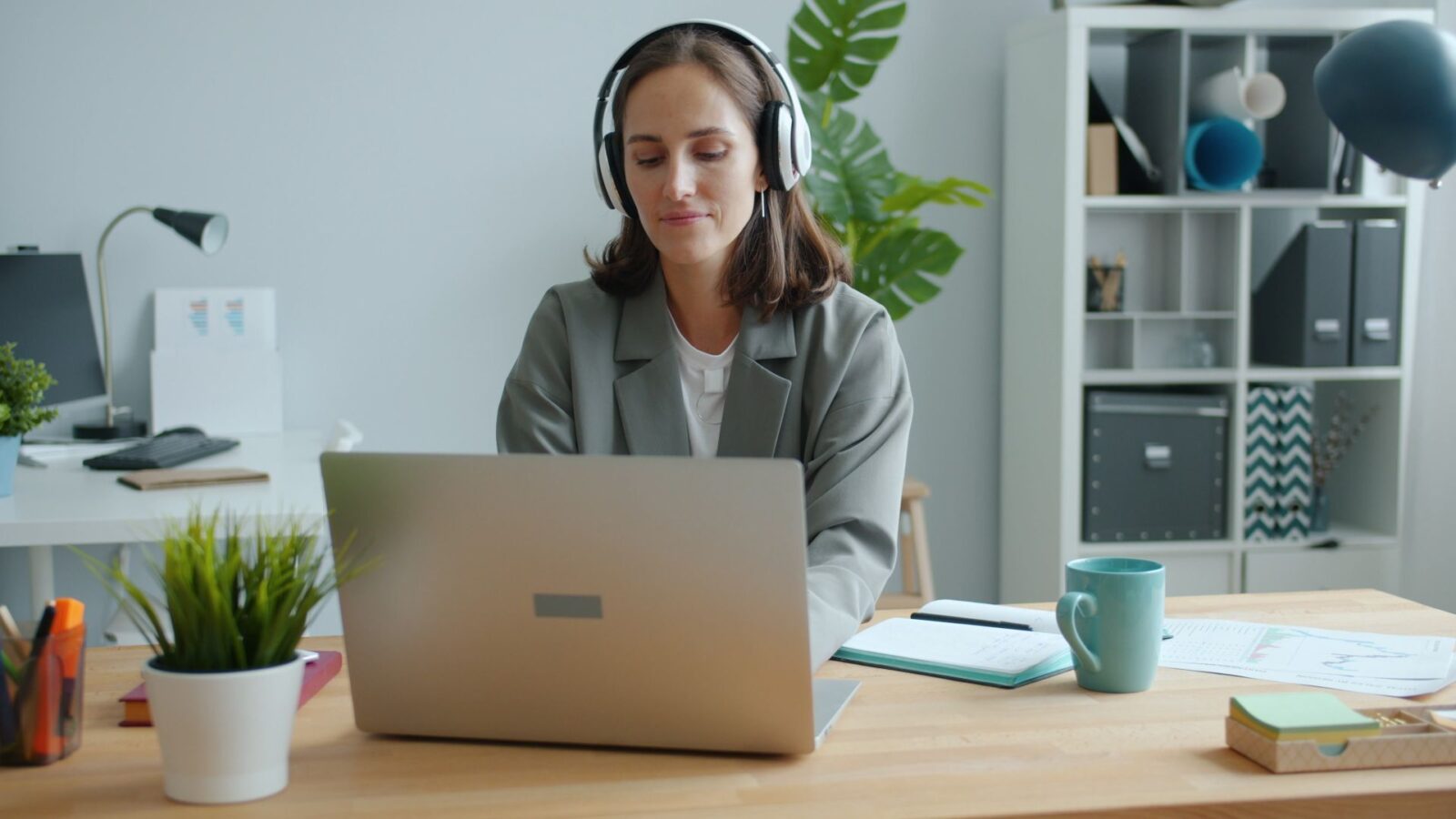 Donna con cuffie seduta a una scrivania davanti a un computer portatile, con quaderno, tazza e piantina accanto