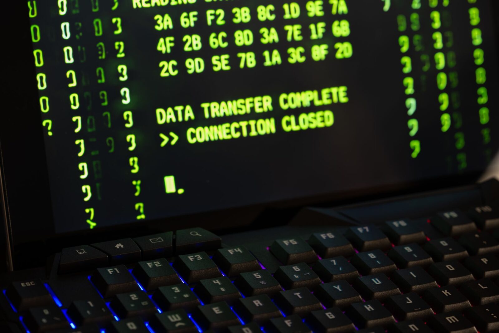 Schermo nero con codice verde e le scritte DATA TRANSFER COMPLETE e >> CONNECTION CLOSED, davanti una tastiera illuminata