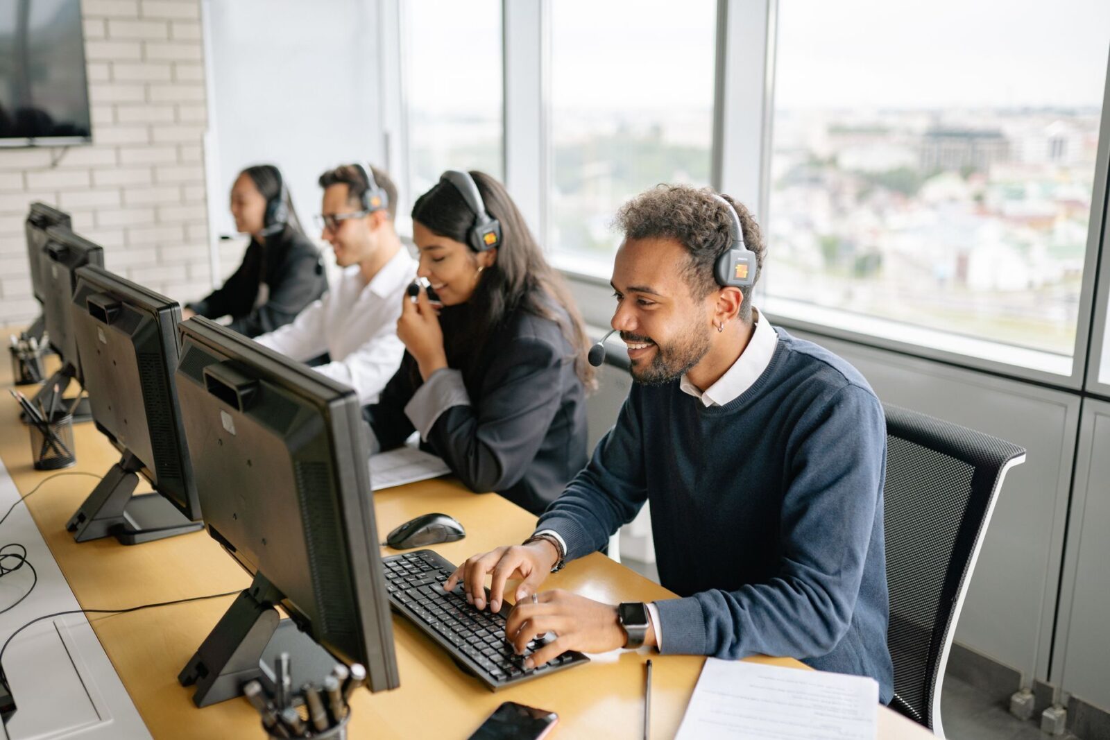Quattro persone con cuffie e microfono lavorano a postazioni con monitor allineate su una scrivania accanto a grandi finestre